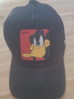 Svart truckerkeps Daffy Duck Capslab - Svart UV-truckerkeps från Capslab med mesh på baksidan och en stor patch  fram med Daffy Duck från Looney Tunes på röd bakgrund. Justerbar snapback och broderad Capslab-logga bak. Perfekt för dig som gillar tecknade serier och streetwear.