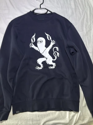 Mörkblå crewneck med ninja-lejon - Säljer en mörkblå crewneck från SILLY med ett stort vitt tryck av ett ninja-lejon på bröstet. Tröjan har rund halsringning, långa ärmar och ribbade muddar. Perfekt för dig som gillar unika prints och streetwear vibes.