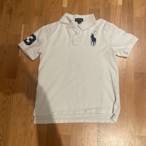 Vit Polo Ralph Lauren pikétröja - Vit big-pony pikétröja från Polo Ralph Lauren med klassisk krage, korta ärmar och stor marinblå logga broderad på bröstet. Snygg detalj med en stor siffra 3 på ena ärmen. Tillverkad i bomull för en skön och luftig känsla. L i ungdom som motsvarar xs. Säljer eftersom den passar inte. Kom med frågor💤