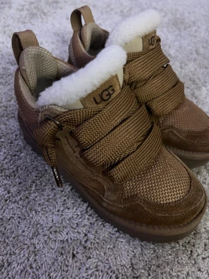 Bruna sneakers från UGG med fårull - Snygga bruna sneakers från UGG med breda snören och detaljer i mocka. Insidan har mjuk vit fårull för extra komfort och värme. Skorna har en chunky sula och UGG-logga på sidan och plösen. Perfekta för dig som vill ha både stil och mysfaktor.