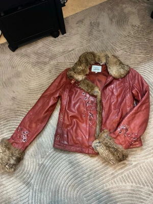 Rosa skinnjacka med fuskpäls Guess - Snygg rosa skinnjacka från Guess by Marciano med broderade blommor på ärmar och framsida. Jackan har bred krage och muddar i brun fuskpäls som ger en unik look. Perfekt för dig som vill sticka ut med en cool och trendig jacka.