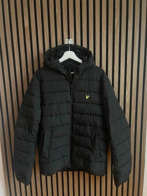 Svart pufferjacka från Lyle & Scott - Svart dunjacka från Lyle & Scott med huva och dragkedja framtill. Jackan har två sidofickor och det klassiska gula logomärket på bröstet. 