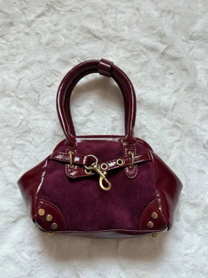 Vinröd handväska med gulddetaljer - Snygg vinröd handväska i en mix av mocka och glansigt skinn. Väskan har två handtag, guldfärgade metalldetaljer och dekorativ spänne framtill. Insidan är fodrad i svart tyg och öppningen har metallram för extra form. Perfekt accessoar för en trendig look.