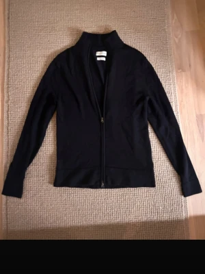 Svart merinoullskofta These Glory Days - Stilren svart kofta från These Glory Days i mjuk superior merinoull. Koftan har hel dragkedja framtill, hög krage och långa ärmar. Perfekt för dig som gillar minimalistisk och klassisk stil.