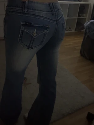 Blå bootcut jeans med fickdetaljer - Säljer ett par blå bootcut jeans med snygga slitningar och kontrastsömmar. Jeansen har dekorativa fickor bak med knappdetalj och låg midja. Perfekta för dig som gillar en avslappnad men trendig look.