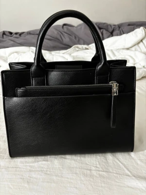 Svart handväska i skinn  - Snygg svart handväska i slätt skinn med två rejäla handtag och silverfärgade dragkedjor. Väskan har en stilren, fyrkantig form och diskret logga framtill. Perfekt för dig som gillar minimalistisk och klassisk design.