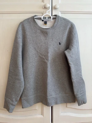Grå sweatshirt från Ralph Lauren - Klassisk grå sweatshirt från Ralph Lauren med rund halsringning och diskret mörkblå logga broderad på bröstet. Tröjan har långa ärmar och mjukt material på insidan, perfekt för en avslappnad och stilren look.