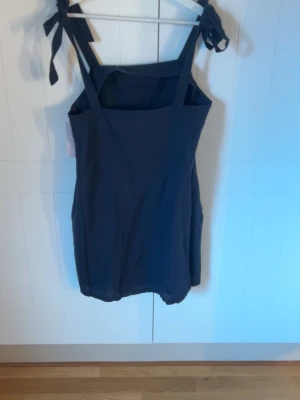 Mörkblå kort klänning med knytband från Nelly - En stilren mörkblå kort klänning med raka linjer och breda axelband som knyts på axlarna. Klänningen har en enkel design och är tillverkad i ett lätt tyg, perfekt för varma dagar. Passar dig som gillar minimalistisk och trendig stil. Liten i storleken 