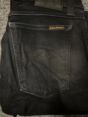 Svarta Nudie Jeans - Säljer ett par svarta skinny jeans från Nudie Jeans med klassisk femficksmodell och diskret vågmönster på bakfickan. Jeansen har silverfärgade nitar och är tillverkade i stretchig denim för en tajt passform. Perfekta för dig som gillar stilrena och bekväma jeans.