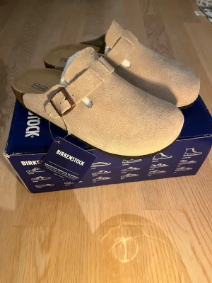 Beige Birkenstock tofflor i mocka - Snygga beige tofflor från Birkenstock i mjukt mockamaterial med justerbart spänne i bronsfärg. Perfekta för att glida runt hemma eller på stan. Klassisk design med bekväm fotbädd och robust sula. Kommer i originalkartong.