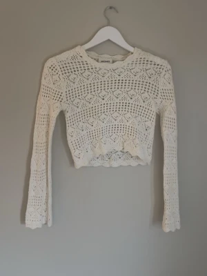Vit virkad cropped tröja Monki - Säljer en vit, virkad cropped stickad tröja från Monki. Tröjan har ett genombrutet mönster, långa ärmar och en vågig kant nedtill. Perfekt för lager på lager och ger en trendig boho-vibe.