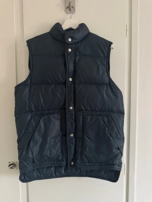 Mörkblå dunväst med hög krage - Snygg mörkblå dunväst med hög krage och tryckknappar framtill. Västen har två stora framfickor och quiltad design som ger en puffig look. Okänt märke.