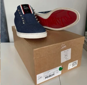 Blå Christian Louboutin sneakers - Säljer ett par blå denim sneakers från Christian Louboutin med ikonisk röd sula och coola silverfärgade nitar längs snörningen. Skorna har vit platt sula och detaljer i vitt och rött på plösen. Insidan är fodrad med perforerat vitt skinn för extra komfort. Fint skick alla nitar kvar, kvitto finns och box o allt. Svåra att få tag på i Sverige!