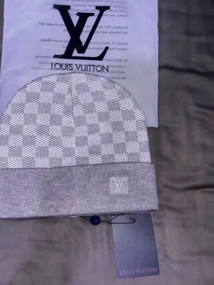 Grå Louis Vuitton mössa med rutmönster - Snygg grå mössa från Louis Vuitton med klassiskt rutmönster och vikt kant. LV-logga broderad framtill på kanten. Tillverkad i mjukt stickat material som känns lyxigt och bekvämt. Perfekt accessoar för att lyfta din outfit.