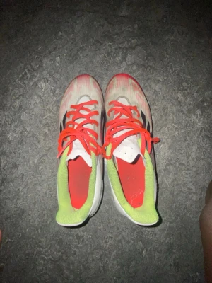 Adidas löparskor med neonröda detaljer - Säljer ett par Adidas löparskor i vitt med neonröda och limegröna detaljer. Skorna har svarta ränder på sidan, röda snören och en sula med rött mönster för extra grepp. Perfekta för löpning eller träning utomhus.