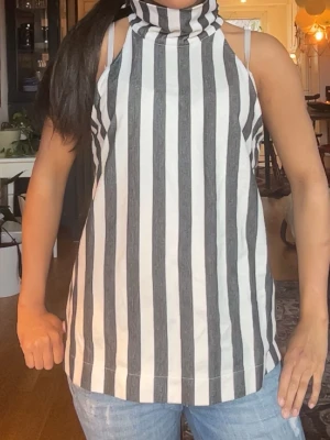 Randig polotopp i svart och vitt - Säljer en ärmlös polotopp med breda svarta och vita ränder. Toppen har hög krage och rak passform, perfekt för en trendig och avslappnad stil. Den här knappar i nacken !