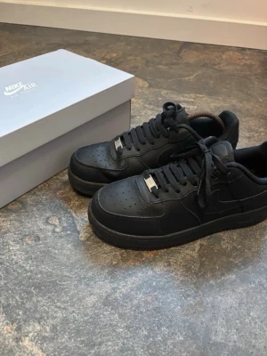Svarta Nike Air Force 1 sneakers - Klassiska helsvarta Nike Air Force 1 sneakers i läder med perforerad tå, svart sula och svarta skosnören. Ikonisk siluett med låg profil och diskret Nike-logga på sidan. Perfekta för en clean och stilren look.
