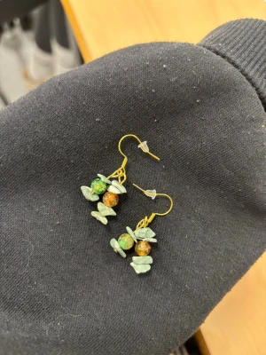 Gröna örhängen med pärlor - Unika örhängen med krok i guldfärg och dekorativa pärlor i grönt och orange. Örhängena har även små gröna stenar i oregelbunden form som ger en bohemisk känsla. Perfekta för dig som gillar färg och naturinspirerade accessoarer.