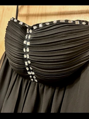 Vero Moda Svart långklänning med strassdetaljer - Elegant svart långklänning med plisserad överdel. Framtill pryds klänningen av glittriga strassdetaljer som ger extra lyxig känsla. Kjolen faller mjukt och materialet har en lätt glans. Perfekt för dig som vill sticka ut. Längd - ca 128cm Byst under - ca 36cm