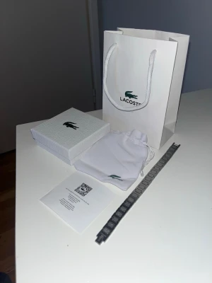 Silverfärgat armband från Lacoste - Snyggt armband från Lacoste i silverfärgad metall med rutigt mönster och diskret Lacoste-logga. Kommer med originalpåse, ask och presentpåse. Perfekt accessoar för dig som gillar stilrena detaljer och vill ha något som sticker ut lite extra.