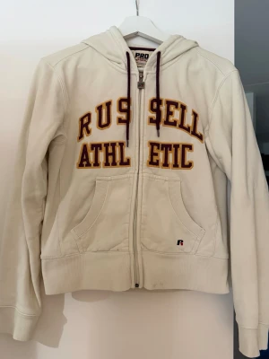 Vit hoodie  Russell Athletic - Vit hoodie med dragkedja från Russell Athletic. Broderad text i vinrött och gult framtill, två fickor och huva med vinröda snören. Sitter mer tajt. Superskön ⭐️ använd några gånger, fläckar går att ta bort