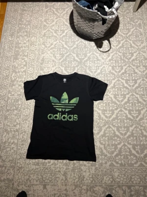 Svart Adidas t-shirt med logga - Svart t-shirt från Adidas med den klassiska gröna trefoil-loggan och texten 'adidas' på bröstet. T-shirten har rund hals, korta ärmar och är gjord i 100% bomull för en skön och avslappnad stil.