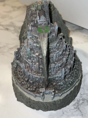 Miniatyr av Minas Tirith - Detaljerad miniatyr av Minas Tirith från Sagan om Ringen. Modellen är gjord i plast och målad i grå, brun och grön färg. Den visar stadens olika nivåer och byggnader med en bergsliknande bas. Perfekt för samlare eller fans av fantasy.