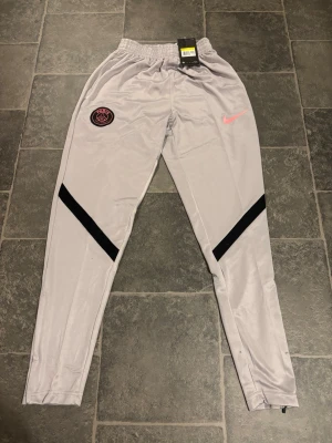 Grå PSG träningsbyxor från Nike - Säljer ett par grå träningsbyxor från Nike med PSG-logga på vänster lår och rosa Nike Swoosh på höger sida. Byxorna har svarta detaljer på benen och elastisk midja. Perfekta för fotbollsträning eller chill. Materialet är lätt och andas.