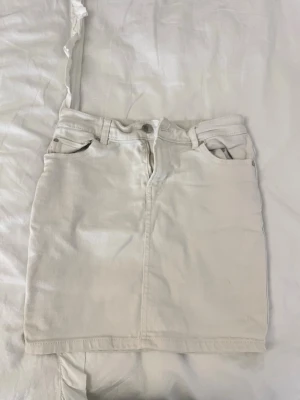 Vit kort jeanskjol med fickor - Snygg vit kort jeanskjol med klassiska fem fickor, bälteshällor och dragkedja framtill. Kjolen har en rak passform och är perfekt för en trendig och avslappnad stil. Passar till allt från sneakers till boots.