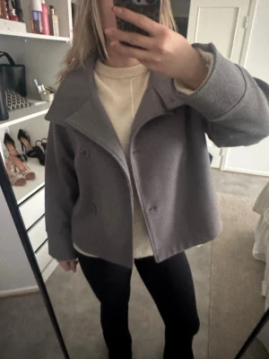 Kort grå ullrock med bred krage - Snygg kort grå ullrock med dubbelknäppning och bred krage. Rocken har en boxig passform och är tillverkad i ett tjockare material som ger en stilren look. Perfekt att slänga över en stickad tröja för en trendig outfit.