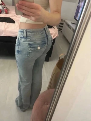 Ljusblå bootcut jeans med fickdetaljer - Säljer ett par ljusblå bootcut jeans med snygga sömmar och fickor med lock och knapp bak. Jeansen har en klassisk femficksmodell och är tillverkade i denim med lätt tvättad look. Perfekta för en avslappnad och trendig stil.