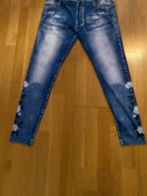 Blå jeans med vita blommor - Snygga blå jeansleggings tights med slitningar och vita blommor tryckta längs benen. Jeansen har en klassisk femficksmodell och smal passform. Perfekta för dig som vill ha något unikt och trendigt i garderoben.