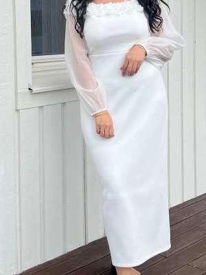 Vit långklänning med blommig kant - Elegant vit långklänning med offshoulder-design och transparenta långärmar. Klänningen har en vacker blommig kant upptill och en figurnära passform. Perfekt för speciella tillfällen och studentfirande.