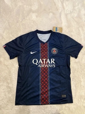 PSG matchtröja Nike blå röd - Snygg PSG matchtröja från Nike i mörkblått med röd och vit detalj i mitten, klubbmärke och Qatar Airways-tryck på bröstet. Tillverkad i lätt och ventilerande polyester, perfekt för fotboll eller träning. Kort ärm och klassisk passform.