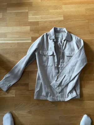 Ljusgrå overshirt med fickor - Stilren ljusgrå overshirt i mjukt tyg med klassisk krage och två stora bröstfickor med lock och knappar. Långa ärmar med manschett och knappar. Perfekt att bära öppen över en t-shirt för en avslappnad look.