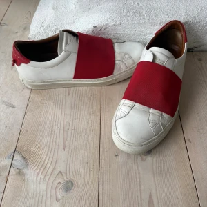 Givenchy urban street sneakers - Säljer ett par vita sneakers från Givenchy med bred röd elastisk rem över foten och röd häl. Skorna har en klassisk låg profil, rund tå och vit sula. Insidan är fodrad i brunt skinn för extra komfort. Perfekta för en stilren och modern look.