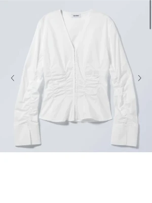 Vit blus med rynk och hakar från Weekday - Stilren vit blus från Weekday med långa ärmar, markerade manschetter och rynkdetaljer längs sidorna. Blusen har en unik knäppning framtill med hakar och är tillverkad i ett glansigt tyg som ger en modern look. Aldrig använd med prislappen kvar - originalpris: 500 kr