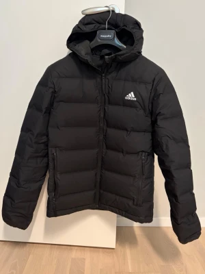 Svart dunjacka från Adidas - Säljer en svart pufferjacka från Adidas med huva och dragkedja framtill. Jackan har två sidofickor med dragkedja och Adidas-logga på bröstet. Den högra fickans dragkedja är trög och fungerar inte helt som den ska. 
