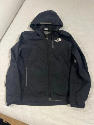 Svart vindjacka från The North Face - Snygg svart vindjacka från The North Face med huva och dragkedja framtill. Jackan har två fickor med dragkedja och logga på bröstet. Perfekt för blåsiga dagar och enkel att matcha med olika outfits.