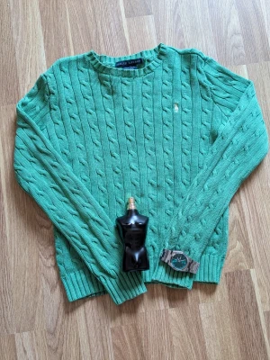 Grön kabelstickad tröja Ralph Lauren - Snygg grön kabelstickad tröja från Ralph Lauren med rund halsringning och diskret broderad logga på bröstet. Tröjan har långa ärmar och ribbade muddar vid ärmslut och nederkant. Perfekt för en chill och stilren look.
