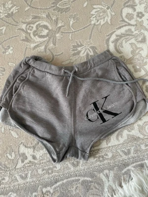  Calvin Klein Jeans shorts  - Snygga grå shorts från Calvin Klein Jeans med snörning i midjan och stort CK-tryck på ena benet. Tillverkade i mjuk bomullsmix, perfekta för chill dagar eller träning. Klassisk passform med fickor på sidorna och elastisk midja.