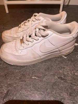 Vita Nike Air Force 1  - Klassiska vita Nike Air Force 1 sneakers i lågt utförande. Skorna har en stilren design med perforerad tå, vita skosnören och den ikoniska swoosh-loggan på sidan. Tillverkade i läder med en robust gummisula för extra komfort.
