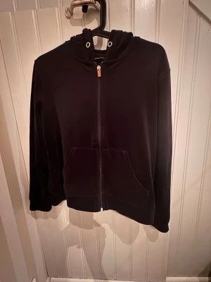 Svart velourhoodie från Gina Tricot - Mysig svart hoodie i velour från Gina Tricot med dragkedja framtill, två stora fickor och huva med metallöljetter. Perfekt för en chill och avslappnad stil. Passar till allt och är superbekväm att bära.