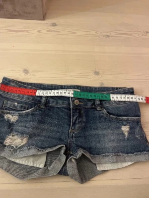 Mörkblå slitna jeansshorts - Säljer ett par mörkblå jeansshorts med slitna detaljer och två knappar i midjan. Shortsen har klassisk femficksdesign, knappgylf och bälteshällor. Perfekta för en avslappnad och trendig look.