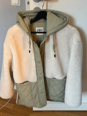 SÖKER ZARA JACKA!! - Den har: vit sherpa/fleece på överdelen och ärmarna grön quiltad nedre del huva tryckknappar framtill Kallas: “ZARA Hooded Sherpa Quilted Jacket” 