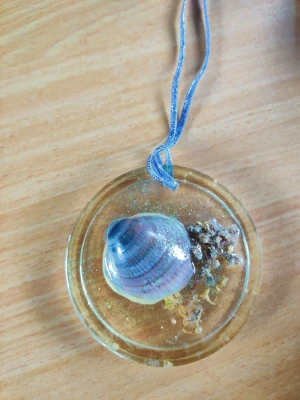 Rund hängande solfångare dekoration med snäcka - Handgjort runt smycke i transparent resin med en skimrande snäcka och torkad växt inuti. Smycket har inslag av glitter och hängs i ett blått band. Perfekt för dig som gillar strandvibbar och unika accessoarer.