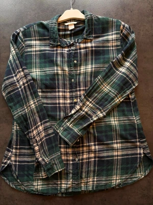 Grönrutig skjorta från H&M - Grön och vitrutig flanellskjorta från H&M.  Skjortan har långa ärmar, knappar framtill och en avslappnad passform. Perfekt att bära öppen över en t-shirt eller stängd för en mer klassisk look. Dammodell så uppsydd i nederkant. 