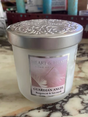 Doftljus Heart & Home Guardian Angel - Säljer ett doftljus från Heart & Home i doften Guardian Angel med toner av bergamott och söt mysk. Ljuset kommer i en frostad glasburk med dekorativt silverfärgat lock och har en enkel veke. Perfekt för att skapa mysig stämning hemma.