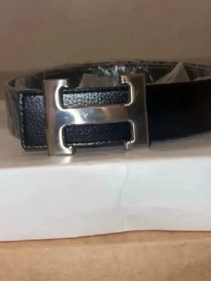 Svart läderbälte med H-spänne - Snyggt svart bälte i läder med ett stort, silverfärgat H-format spänne. Perfekt accessoar för att lyfta vilken outfit som helst och ger en clean och stilren look. Passar till jeans eller kostymbyxor.