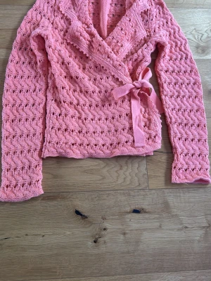 Rosa omlottkofta från Odd Molly - (SAMPLE)  Säljer en stickad rosa omlottkofta från Odd Molly med knytband i midjan och ett fint hålmönster. Koftan har långa ärmar och bred krage, perfekt för lager på lager. Materialet är mjukt och koftan har en feminin look.